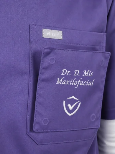 Purple medical scrubs pocket with embroidered text 'Dr. D. Mis Maxilofacial' and logo, showing brand tag near top-left of pocket.