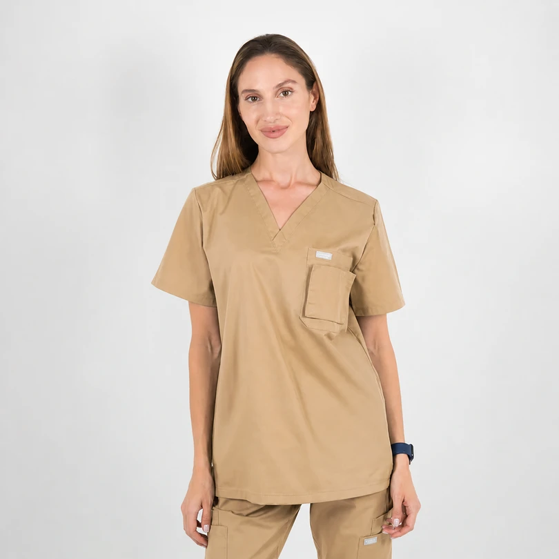 Woman wearing tan medical scrubs with a chest pocket, standing against a light background.