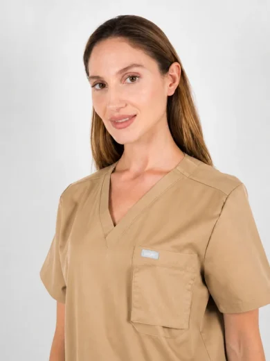 Female healthcare worker wearing a beige scrub top with a chest pocket, facing slightly to the right against a light background.