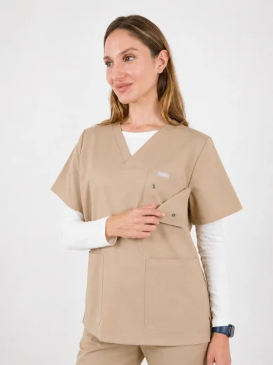 Female healthcare worker wearing beige scrubs and a white undershirt, smiling and looking to the side with one hand resting on the pocket.