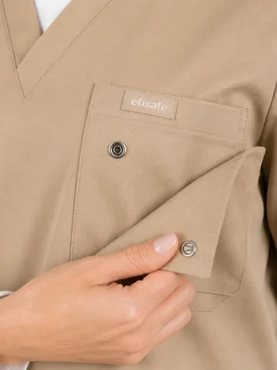 Beige medical scrubs top with a front pocket and a hand adjusting a snap-button flap, brand label visible above pocket.