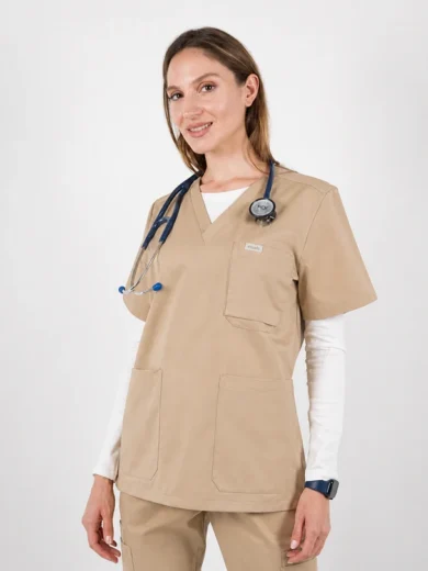 Female healthcare professional in beige scrubs with stethoscope around neck, smiling at camera in a clinical setting, wearing white underlayer and smartwatch.