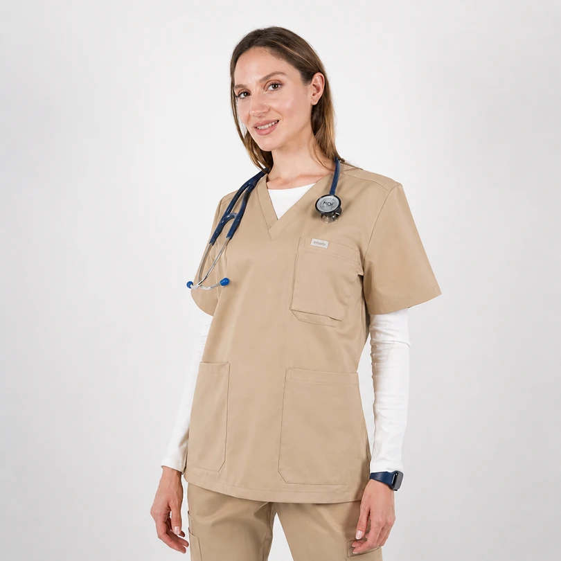 Female healthcare professional in beige scrubs with stethoscope around neck, smiling at camera in a clinical setting, wearing white underlayer and smartwatch.
