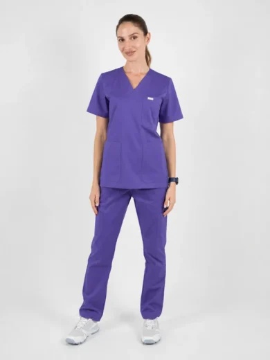Female healthcare worker wearing purple scrubs (short-sleeve top with pockets and matching pants) standing against a light background, white sneakers.