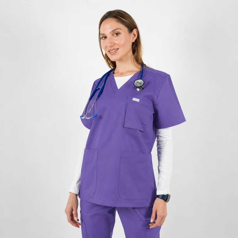Female healthcare worker in purple scrubs with stethoscope around her neck, standing and smiling.