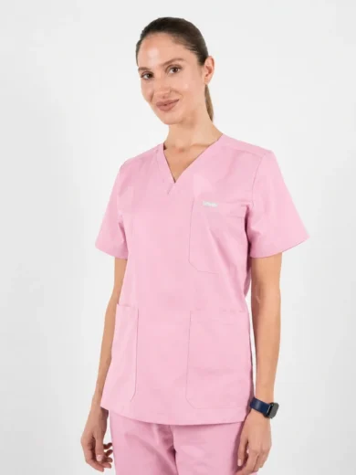 Woman wearing light pink medical scrubs (short-sleeve top with chest and two lower pockets) standing against a white background, smiling subtly.