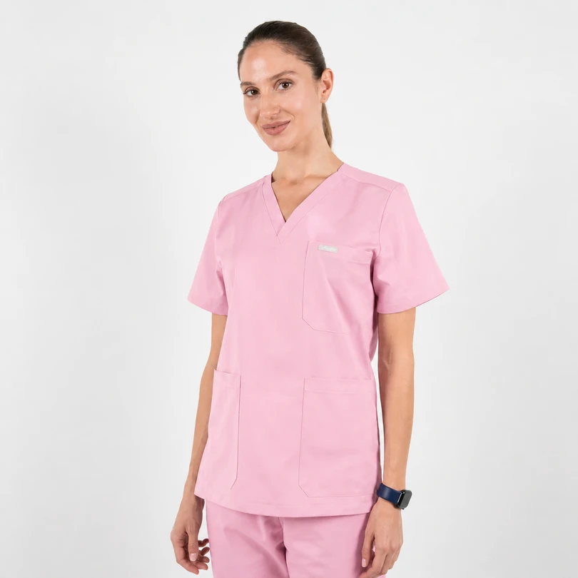 Woman wearing light pink medical scrubs (short-sleeve top with chest and two lower pockets) standing against a white background, smiling subtly.