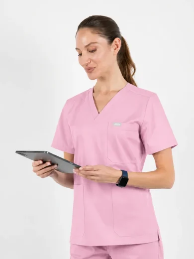 Healthcare worker in pink scrubs standing against a white background, holding and looking at a tablet.