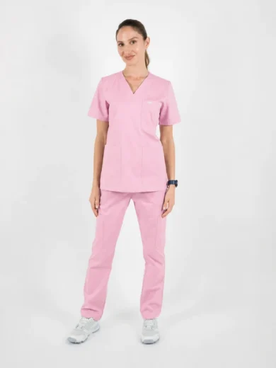 Female model wearing light pink medical scrubs (top with V-neck and large pockets, matching pants) standing in a neutral studio setting, wearing white sneakers and a dark watch on one wrist.