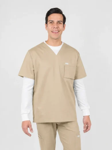 Male healthcare worker wearing beige scrubs with a white undershirt, smiling at the camera, standing in a neutral space.