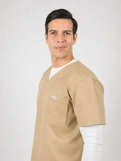 Male healthcare worker in tan scrubs, standing against a light background and smiling at the camera