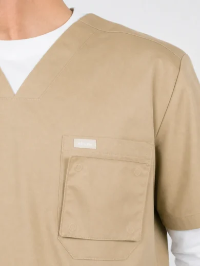 Beige medical scrub top with a V-neck and a chest pocket featuring a small brand label.