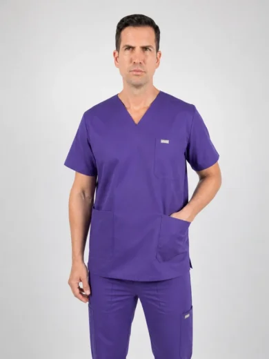 Male healthcare professional wearing purple scrubs with V-neck, standing with hands in pockets against a light gray background.