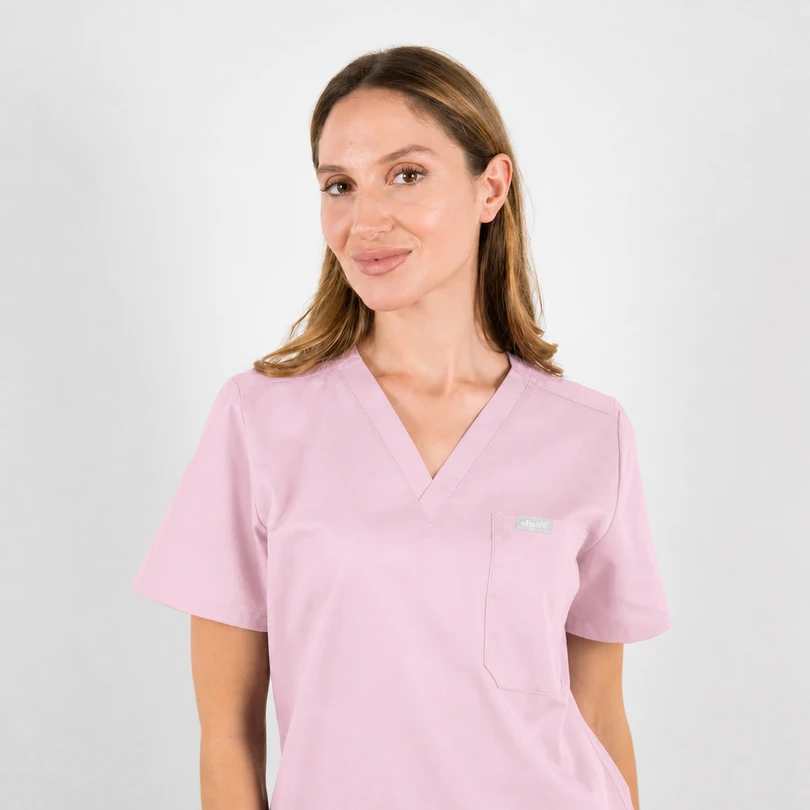 Woman wearing light pink scrubs with V-neck, facing slightly to the left, smiling against a neutral background.