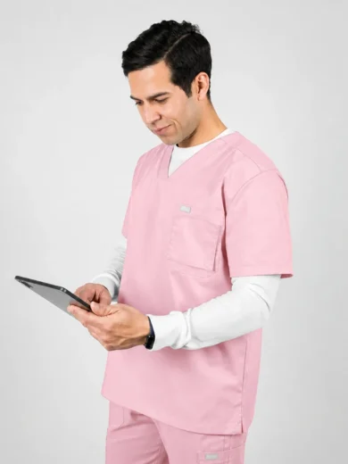 Male healthcare professional in pink scrubs using a tablet while standing against a light gray background, looking at the screen.