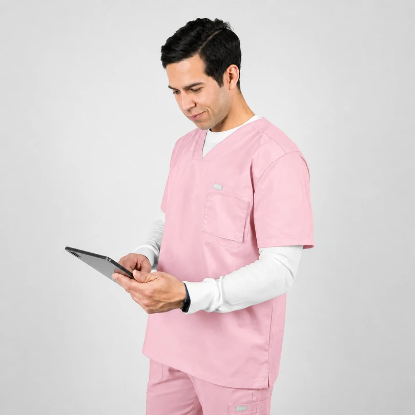 Male healthcare professional in pink scrubs using a tablet while standing against a light gray background, looking at the screen.