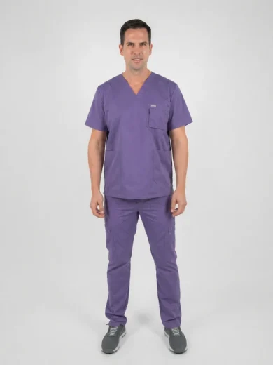 Male healthcare professional standing and facing camera in purple scrubs with short sleeves and pockets on top and pants, neutral studio backdrop.