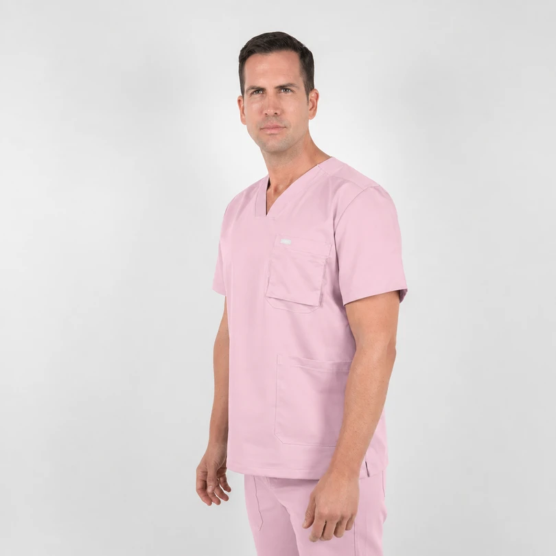 Man wearing light pink medical scrubs standing in a studio, facing slightly to the right.