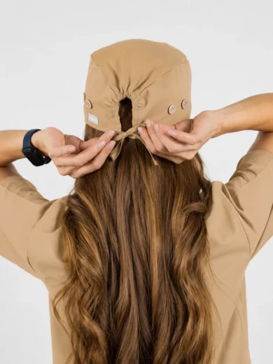 Person with long brown hair wearing a beige cap from the back, adjusting the strap at the back of the cap.