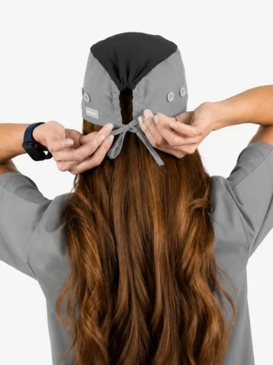 Person photographed from behind tying a gray medical cap over long brown hair while wearing gray scrubs and a watch on the left wrist