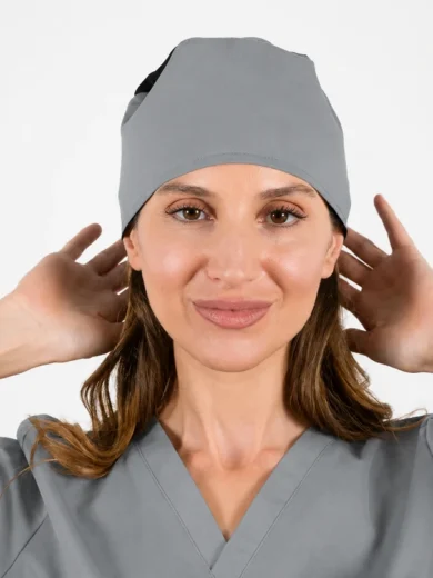 Female healthcare worker in gray scrubs and cap adjusting her headwear, smiling at the camera