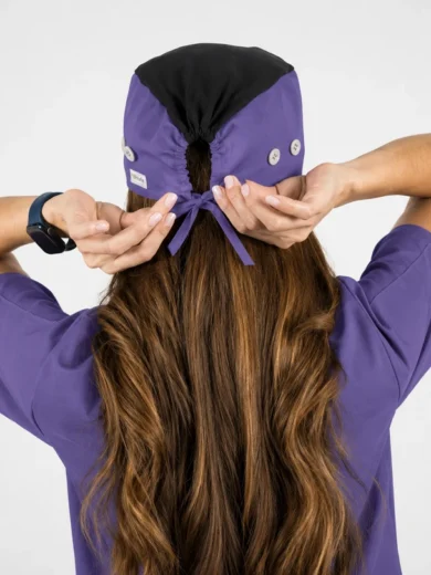 Person in purple scrubs tying a purple scrub cap from behind, long brown hair visible down their back.