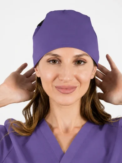 Woman in purple scrubs adjusts a matching scrub cap while smiling at the camera with hands near her ears down to shoulders height