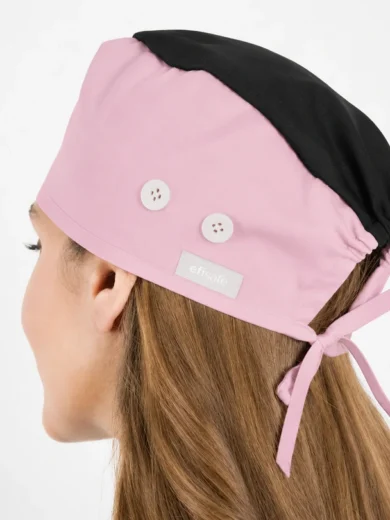 Pink chef hat with black top worn by a person, tied at the back with a bow; two white buttons and a small brand label visible on the side.