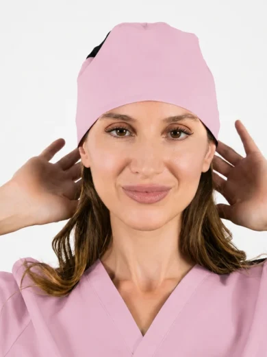 Woman in pink medical scrubs and cap, hands near her ears, smiling at the camera.