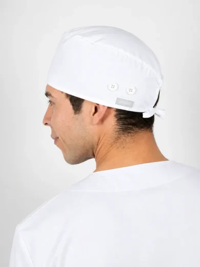 Side profile of a man wearing a white surgical scrub cap with a tie at the back, dressed in white scrubs.