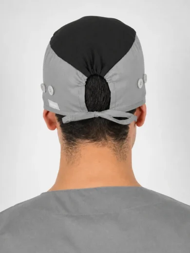 Back view of a person wearing a gray surgical cap with a black top panel, tied at the back with a bow, buttons along the sides, and gray scrubs.