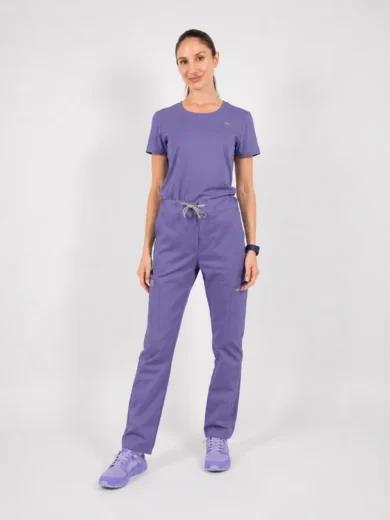 Woman in lavender scrubs (short-sleeve top and drawstring pants) standing with hands by sides, wearing matching lavender sneakers against a light gray background.