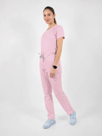 Full-body portrait of a person in light pink medical scrubs, standing in a neutral studio setting, facing slightly to the left.