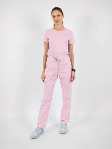 Woman wearing a light pink scrub top and matching drawstring pants, standing in a neutral studio with light blue sneakers.