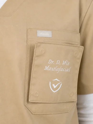 Beige medical scrubs featuring a chest pocket with a removable badge reading 'Dr. D. Mis Maxilofacial' and a logo beneath.
